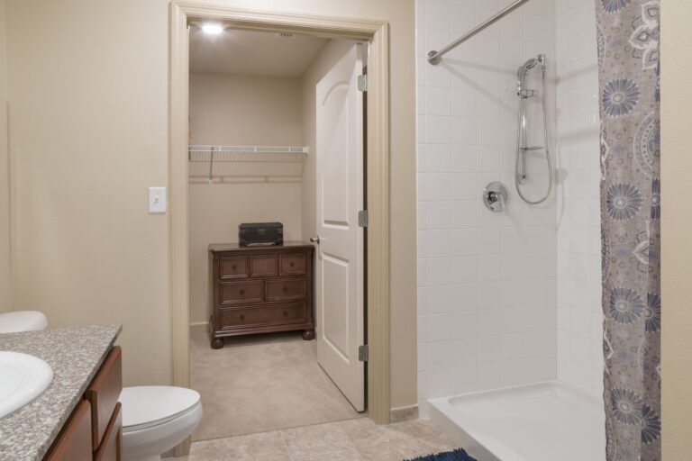 A bathroom with a sink and countertop on the left, a white tiled shower with a curtain on the right, and a doorway leading to a walk-in closet with a dresser and wire shelf.