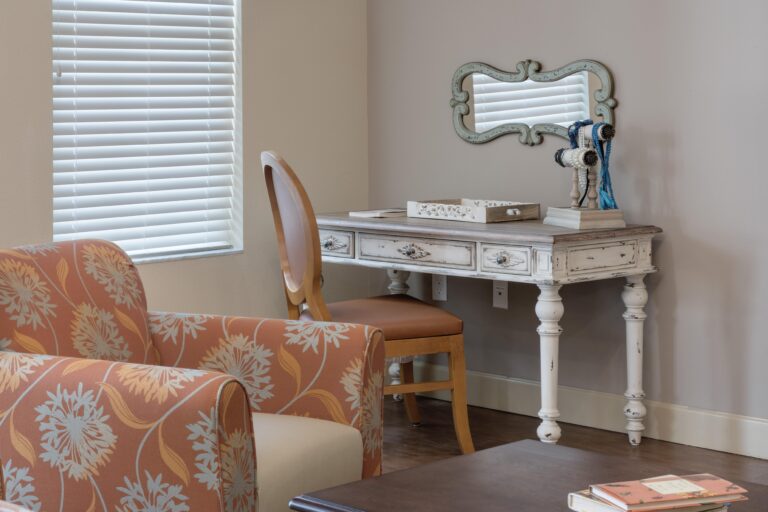 A cozy corner with a floral-patterned armchair, a wooden coffee table, and a vintage white desk with a matching chair. The desk has books, a mirror, and decorative items, next to a window with closed blinds.