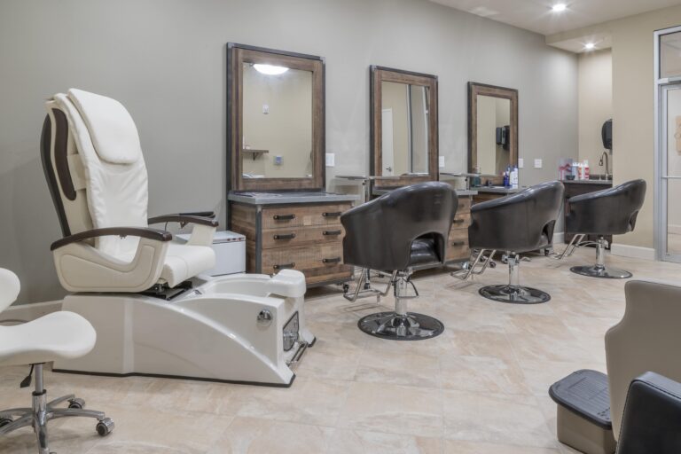 A modern salon interior with a white pedicure chair and three black salon chairs in front of wooden mirrors and cabinets, set against beige walls and a tiled floor.