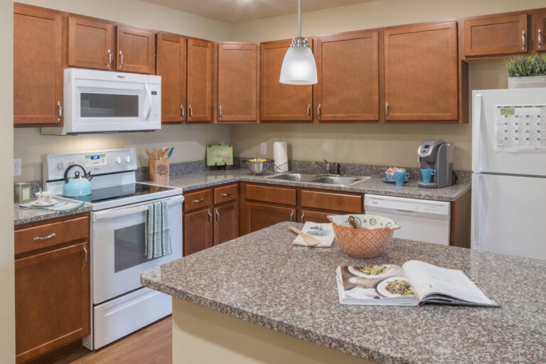 A modern kitchen with brown cabinets, granite countertops, white appliances, a teal kettle on the stove, a coffee maker, a bowl on the counter, and an open cookbook. A pendant light hangs above the island.