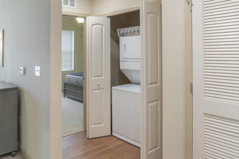 A stacked washer and dryer are tucked in a small laundry closet with double doors in a hallway near a bedroom. The area has light-colored walls and wood flooring.