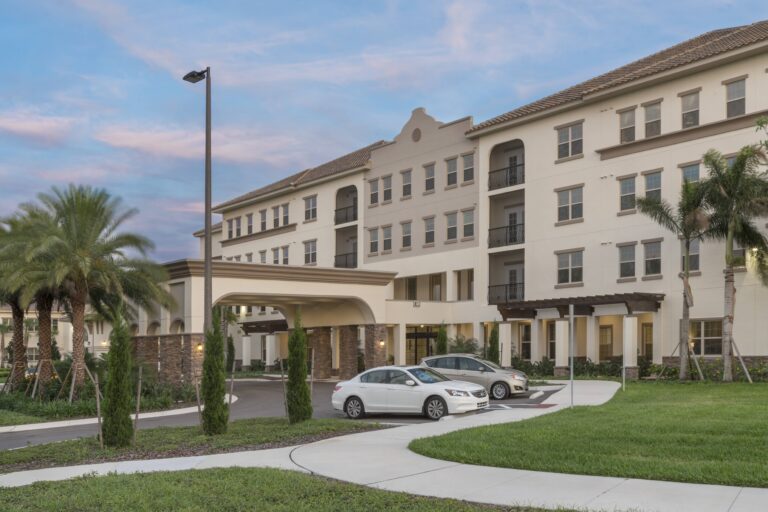 A modern, multi-story residential building with balconies, palm trees, and a covered entrance. Several cars are parked on the driveway, with well-maintained grass and landscaping in the foreground.