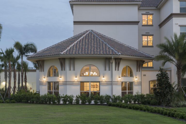 A modern building with a tile roof and large arched windows is surrounded by green grass, palm trees, and shrubs. The windows and exterior lights are warmly lit, creating an inviting appearance at dusk.