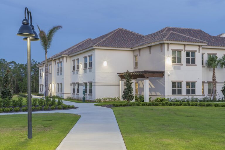 A modern, two-story building with a tile roof is surrounded by manicured lawns, sidewalks, palm trees, and exterior lights at dusk.