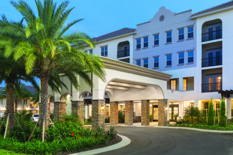 A modern multi-story building with large windows and an arched entrance, surrounded by palm trees, greenery, and a circular driveway, under a clear blue sky at dusk.