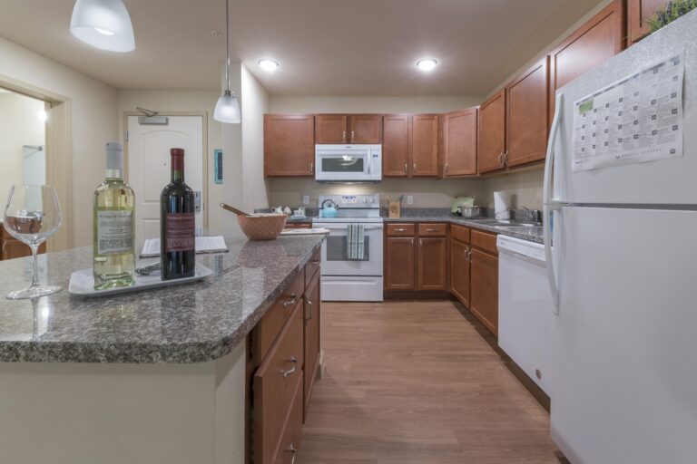 Modern kitchen with wooden cabinets, white appliances, and granite countertops. Two bottles of wine, a wine glass, and a basket are on the counter. The kitchen is well-lit and tidy.