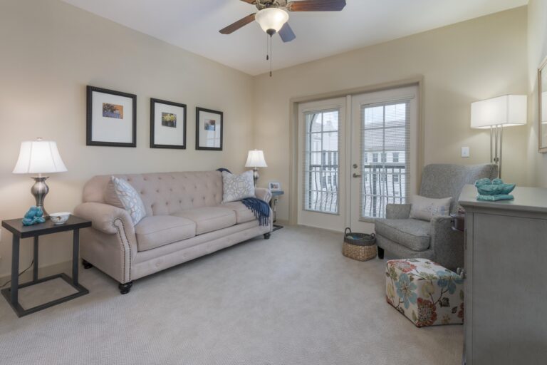 A cozy living room with a beige sofa, armchair, floral ottoman, framed pictures on the wall, lamps, a ceiling fan, and French doors leading to a balcony. The decor is neutral with blue accents.