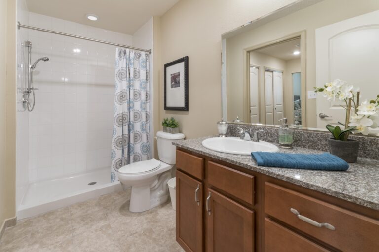 A modern bathroom with a glass-enclosed shower, toilet, granite countertop vanity with a sink, wooden cabinets, a wall mirror, and decorative items such as a plant, towel, and orchids.