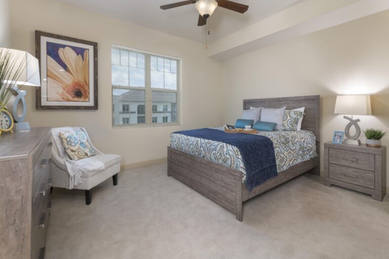 A tidy bedroom with a wooden bed, blue and white bedding, a nightstand with a lamp, a dresser, an upholstered chair, and a large window letting in daylight. A framed flower picture hangs on the wall above the chair.