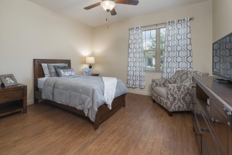 A cozy bedroom with a wooden bed, gray bedding, a patterned armchair, nightstand with lamp, dresser with TV, and a window with geometric curtains letting in natural light.