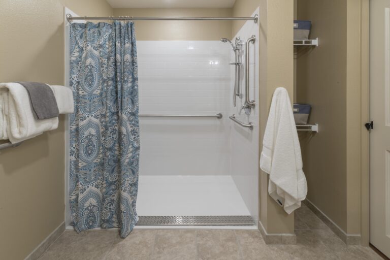 Accessible shower with a blue patterned curtain partially open, grab bars installed on the walls, and a handheld showerhead. White towels hang on racks nearby, and shelves with bins are on the right. Beige tiled floor.