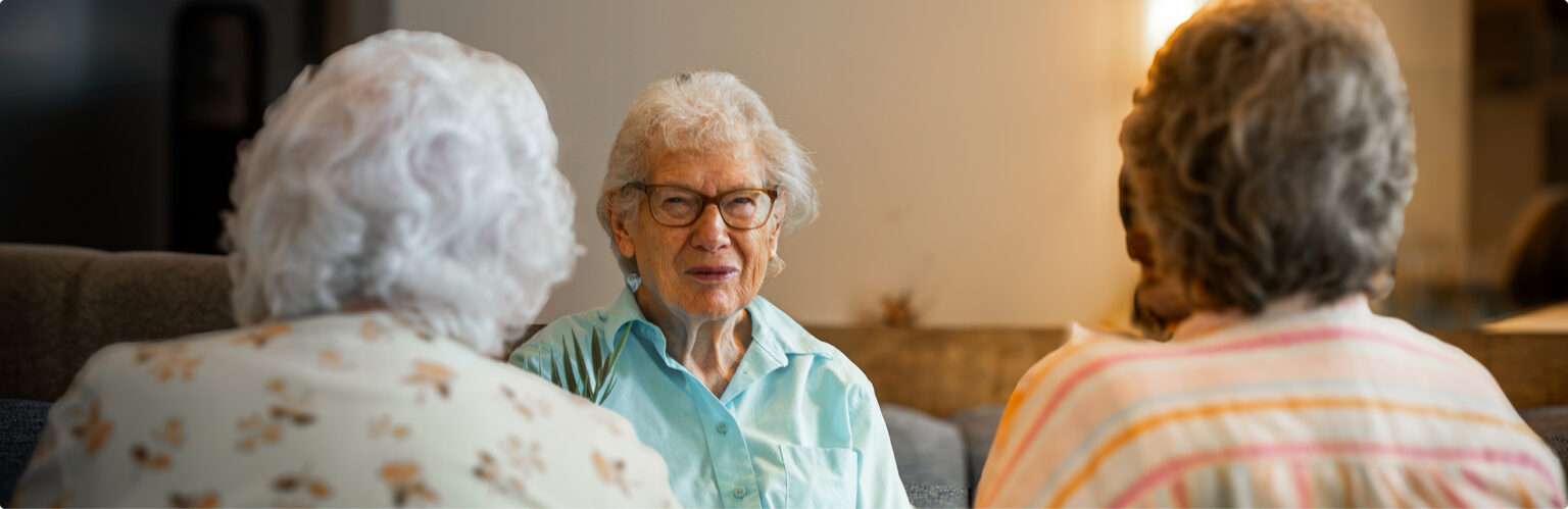 Three senior women sit together indoors, engaged in conversation. The woman in the center, wearing glasses and a light blue shirt, faces the camera, while the other two women are seen from behind.