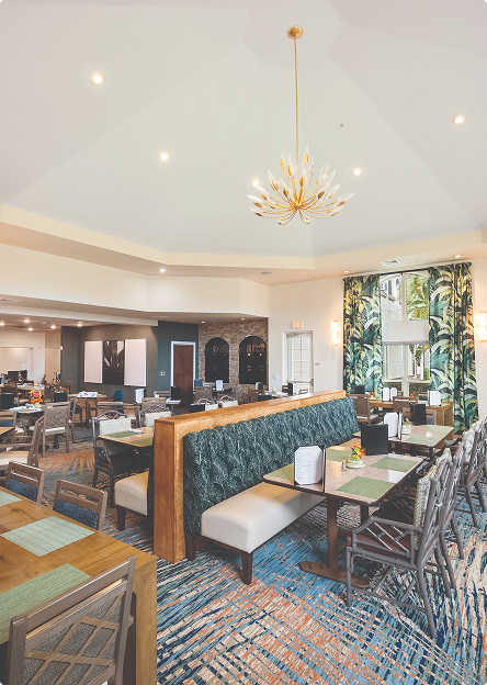A bright, modern dining area with patterned carpet, green cushioned booth seating, wooden tables and chairs, leafy green curtains, and a decorative chandelier hanging from a high ceiling.