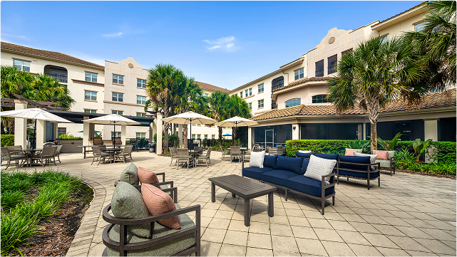 Sunny outdoor patio with cushioned lounge chairs, sofas, tables, and umbrellas surrounded by greenery and palm trees, adjacent to a light-colored multi-story building.