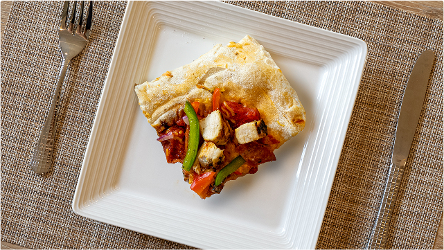 A square slice of pizza topped with grilled chicken, green bell pepper, and tomato on a white square plate, set on a woven placemat with a fork and knife on either side.