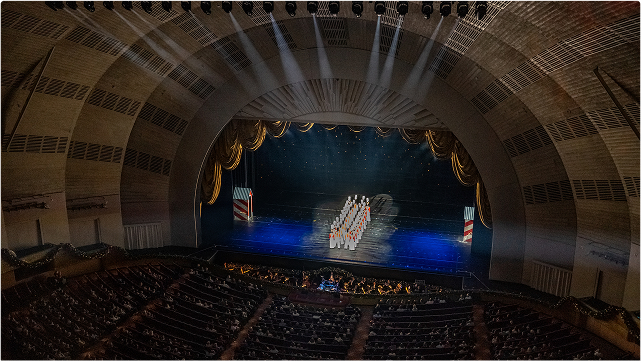 A large theater with an arched ceiling shows a stage performance by a group of dancers in white costumes, standing in formation under bright stage lights, as an audience watches from the seats below.