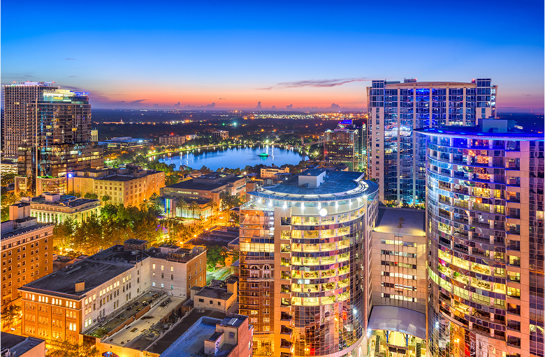 A vibrant cityscape at dusk shows tall, modern buildings with illuminated windows, streets lined with lights, and a lake in the background reflecting the colorful sunset sky.