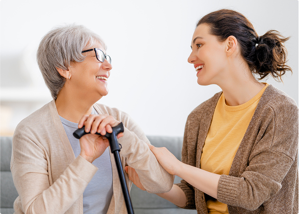 An older woman sits smiling, holding a cane, while a younger woman beside her holds her arm supportively. Both look happy and comfortable in a cozy indoor setting.