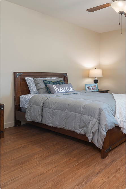A cozy bedroom with a wooden bed frame, gray bedding, pillows including one that says home, a nightstand with a lamp, a framed photo, and wood flooring. A ceiling fan is partially visible above.