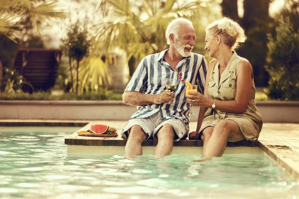 An elderly couple sits on the edge of a pool, smiling and holding drinks. The man wears a striped shirt and shorts; the woman wears a light sleeveless dress. There is a plate with watermelon beside them on a sunny day.