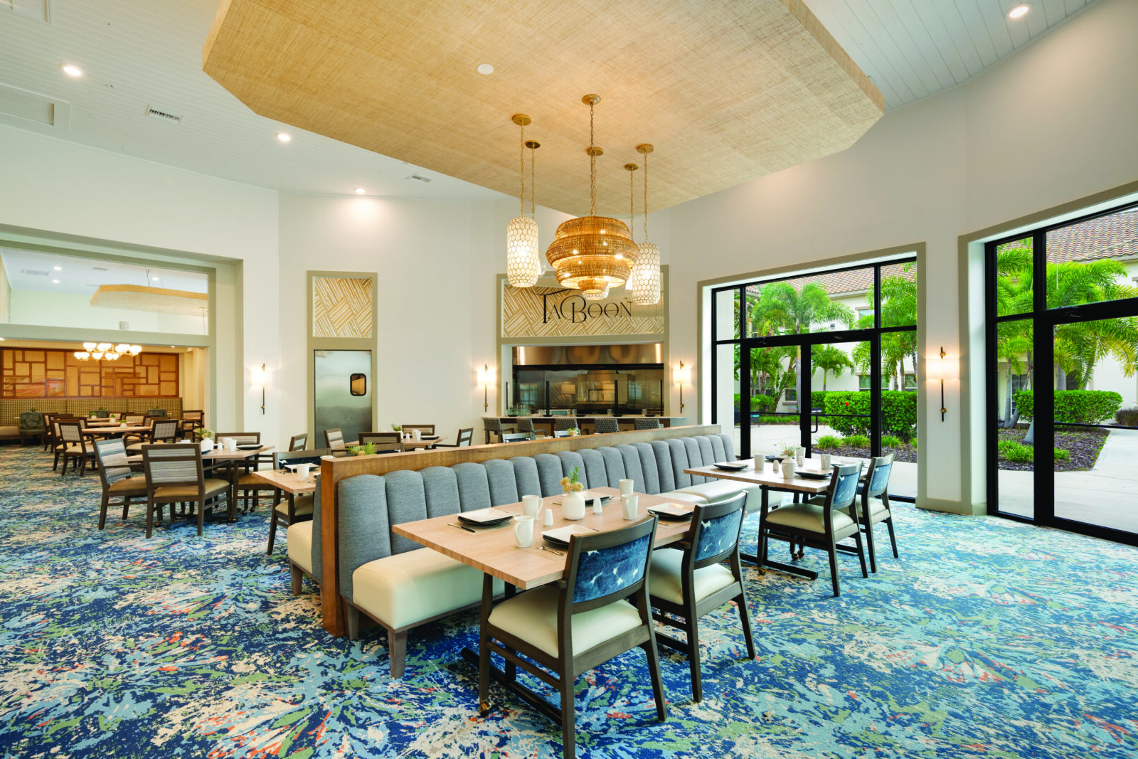 Bright, modern dining area: a long banquette with wooden tables and chairs, patterned blue carpet, large windows overlooking tropical landscaping, and pendant lights hanging from a vaulted ceiling.