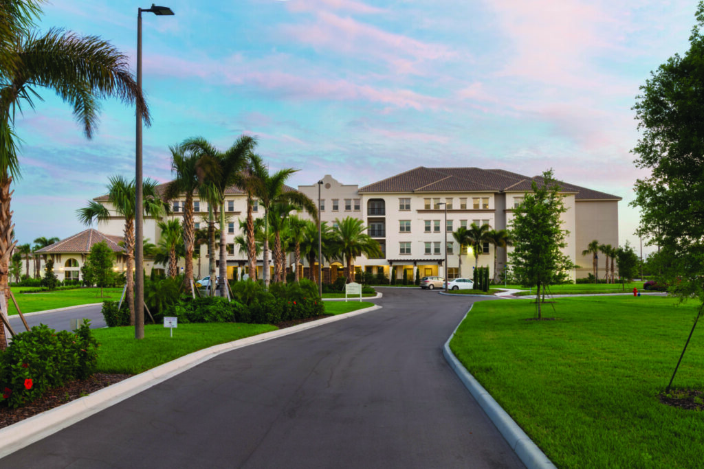 A wide, paved driveway leads to a large, modern building surrounded by palm trees and manicured lawns under a pastel sunset sky.