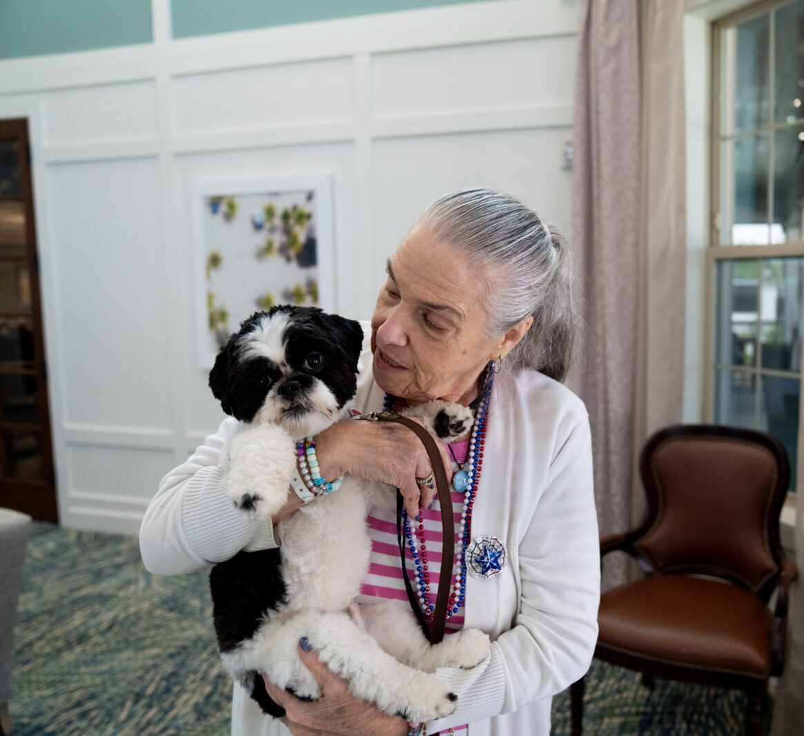 Elderly woman with gray hair in a ponytail smiles warmly while holding a small black-and-white dog in her arms inside a bright community room.