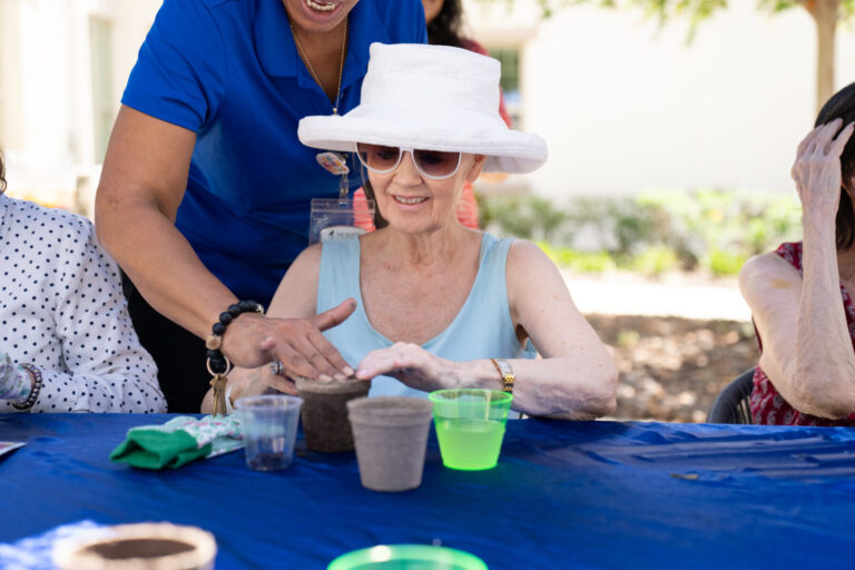 Smiling elderly woman in a sunhat and sunglasses plants seeds in a small pot with the help of a staff member during an outdoor gardening activity.