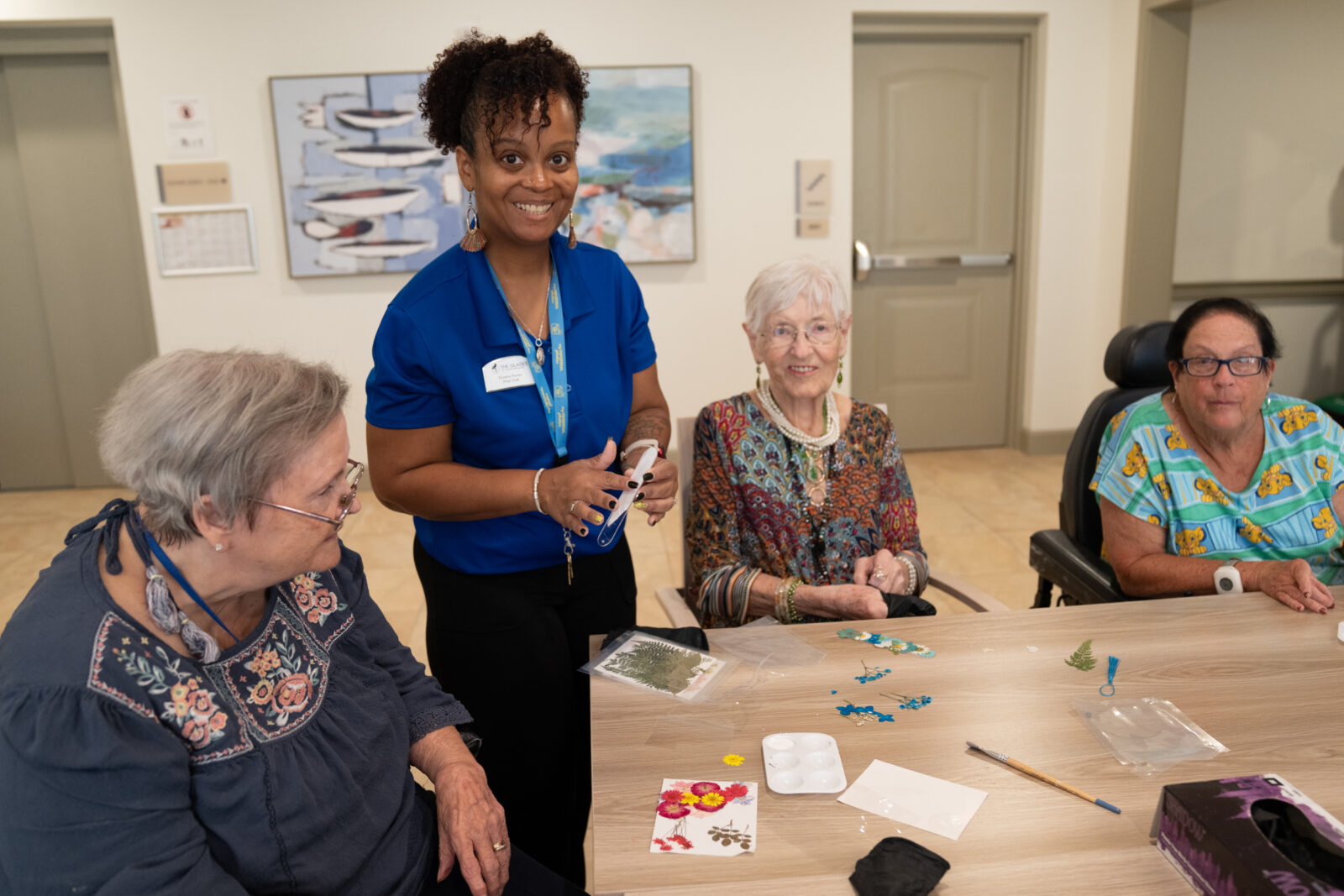 Group of senior women sit at a table working on an arts and crafts project with the help of a smiling staff member in a blue shirt.