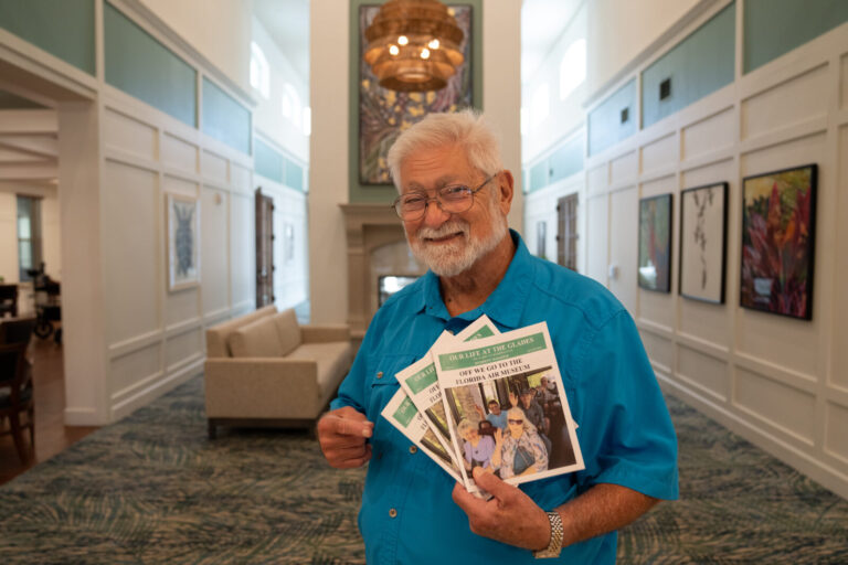 Smiling elderly man with white hair and beard holds community newsletters inside a bright, modern lounge.
