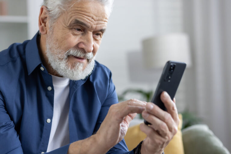 Older man using a smartphone at home, demonstrating safe technology habits to avoid holiday scams.